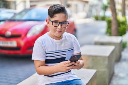 Téléchargez les photos : Adorable garçon hispanique utilisant smartphone assis sur le banc à la rue - en image libre de droit