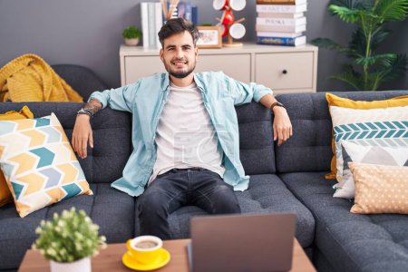 Téléchargez les photos : Jeune homme hispanique souriant confiant assis sur le canapé à la maison - en image libre de droit