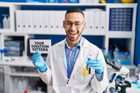 Photo for Young hispanic man working at scientist laboratory holding your donation matters holding blood sample celebrating crazy and amazed for success with open eyes screaming excited. - Royalty Free Image