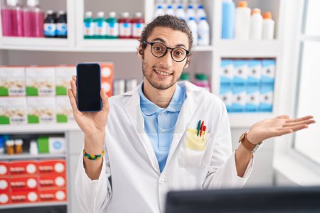 Téléchargez les photos : Jeune homme hispanique travaillant à la pharmacie drugstore montrant écran smartphone célébrant la réalisation avec sourire heureux et expression gagnante avec la main levée - en image libre de droit