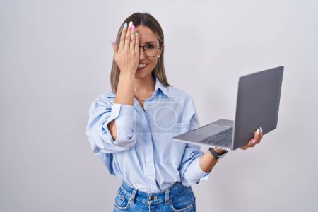 Téléchargez les photos : Jeune femme travaillant à l'aide d'un ordinateur portable couvrant un oeil avec la main, sourire confiant sur le visage et l'émotion surprise. - en image libre de droit