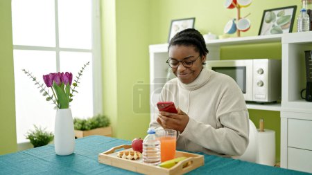 Photo for African american woman having breakfast using smartphone at dinning room - Royalty Free Image