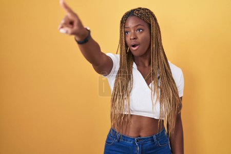 Photo for African american woman with braided hair standing over yellow background pointing with finger surprised ahead, open mouth amazed expression, something on the front - Royalty Free Image