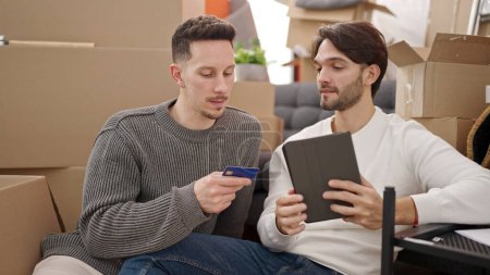 Photo for Two men couple shopping with touchpad and credit card sitting on floor at new home - Royalty Free Image