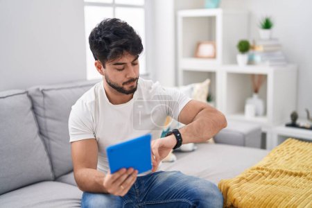 Photo for Hispanic man with beard using touchpad sitting on the sofa looking at the watch time worried, afraid of getting late - Royalty Free Image