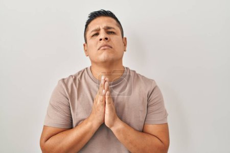 Téléchargez les photos : Jeune homme hispanique debout sur fond blanc mendier et prier avec les mains ainsi que l'expression de l'espoir sur le visage très émotionnel et inquiet. mendier. - en image libre de droit
