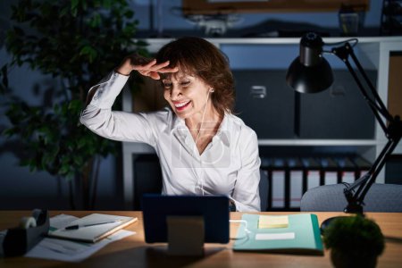 Photo for Middle age woman working at the office at night very happy and smiling looking far away with hand over head. searching concept. - Royalty Free Image