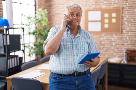 Téléchargez les photos : Moyen âge homme aux cheveux gris travailleur d'affaires en utilisant pavé tactile parler sur smartphone au bureau - en image libre de droit