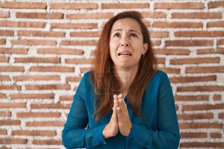 Téléchargez les photos : Femme brune debout sur le mur de briques mendier et prier avec les mains ainsi que l'expression d'espoir sur le visage très émotionnel et inquiet. mendier. - en image libre de droit