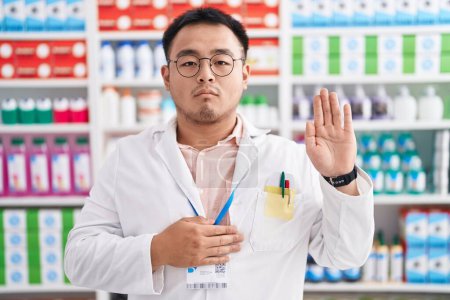 Photo for Chinese young man working at pharmacy drugstore swearing with hand on chest and open palm, making a loyalty promise oath - Royalty Free Image