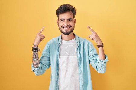 Téléchargez les photos : Young hispanic man with tattoos standing over yellow background smiling pointing to head with both hands finger, great idea or thought, good memory - en image libre de droit