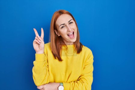 Photo for Young woman standing over blue background smiling with happy face winking at the camera doing victory sign with fingers. number two. - Royalty Free Image