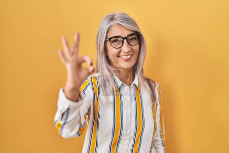 Foto de Mujer de mediana edad con el pelo gris de pie sobre el fondo amarillo con gafas sonriendo positiva haciendo signo de ok con la mano y los dedos. expresión exitosa. - Imagen libre de derechos