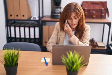 Foto de Middle age woman business worker talking on smartphone using laptop at office - Imagen libre de derechos