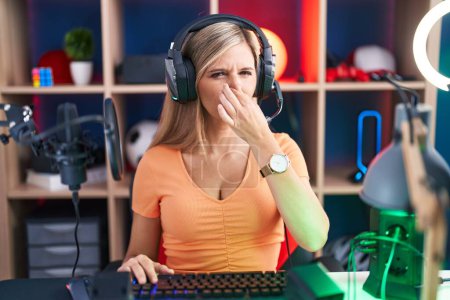 Téléchargez les photos : Jeune femme jouant à des jeux vidéo sentant quelque chose de puant et dégoûtant, odeur intolérable, retenant souffle avec les doigts sur le nez. mauvaise odeur - en image libre de droit