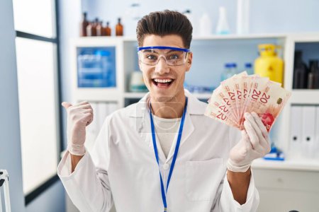 Téléchargez les photos : Jeune homme hispanique travaillant au laboratoire scientifique tenant des shekels pointant le pouce vers le côté souriant heureux avec la bouche ouverte - en image libre de droit