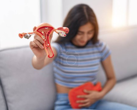 Foto de Chica hispana joven que sufre de dolor menstrual sosteniendo el modelo anatómico del útero en casa - Imagen libre de derechos