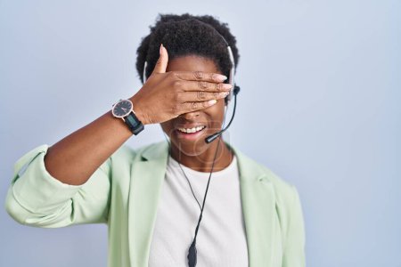 Foto de Mujer afroamericana con auriculares de agente de call center sonriendo y riendo con los ojos tapados de la mano a la cara para sorpresa. concepto ciego. - Imagen libre de derechos