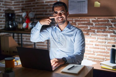 Téléchargez les photos : Homme hispanique avec la barbe travaillant au bureau la nuit pointant du doigt la main vers le visage et le nez, souriant gai. concept de beauté - en image libre de droit