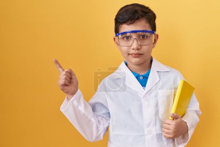 Foto de Pequeño niño hispano con gafas científicas sonriendo feliz señalando con la mano y el dedo hacia un lado - Imagen libre de derechos