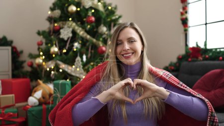 Téléchargez les photos : Young blonde woman doing heart gesture sitting on sofa by christmas tree at home - en image libre de droit
