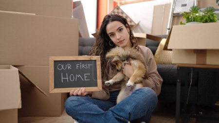 Foto de Mujer hispana joven con perro sosteniendo pizarra sentada en el piso en casa nueva - Imagen libre de derechos