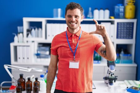 Photo for Young hispanic man working at scientist laboratory smiling and confident gesturing with hand doing small size sign with fingers looking and the camera. measure concept. - Royalty Free Image