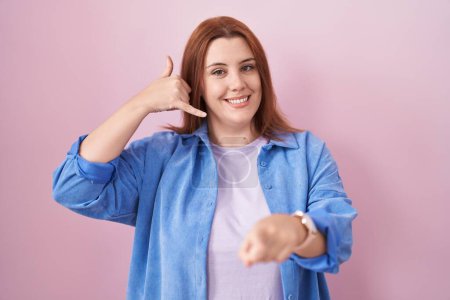 Foto de Mujer hispana joven con el pelo rojo de pie sobre fondo rosa sonriendo haciendo hablar por teléfono gesto y señalando a usted. Llámame.. - Imagen libre de derechos