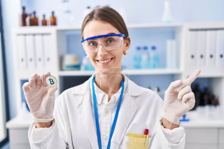 Photo for Young caucasian woman working at scientist laboratory holding bitcoin smiling happy pointing with hand and finger to the side - Royalty Free Image
