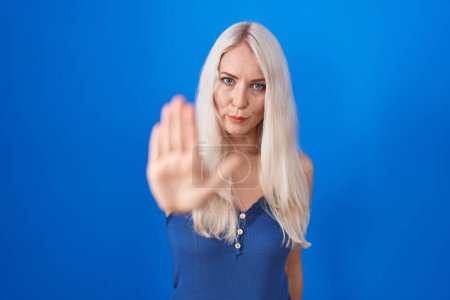 Téléchargez les photos : Femme blanche debout sur fond bleu faire arrêter de chanter avec la paume de la main. expression d'avertissement avec geste négatif et sérieux sur le visage. - en image libre de droit