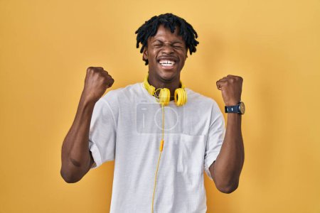 Téléchargez les photos : Jeune homme africain avec dreadlocks debout sur fond jaune célébrant surpris et étonnés pour le succès avec les bras levés et les yeux fermés. concept gagnant. - en image libre de droit
