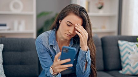 Photo for Young beautiful hispanic woman using smartphone sitting on sofa with sad expression at home - Royalty Free Image