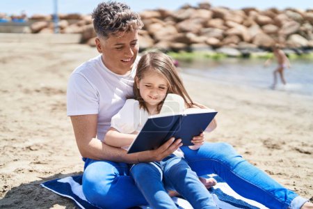 Téléchargez les photos : Father and daughter reading book sitting on sand at beach - en image libre de droit