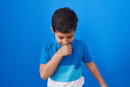 Téléchargez les photos : Petit garçon hispanique debout sur fond bleu se sentant mal à l'aise et toussant comme symptôme de rhume ou de bronchite. concept de soins de santé. - en image libre de droit