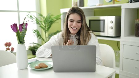 Photo for Young beautiful hispanic woman eating cookies using laptop doing video call at dinning room - Royalty Free Image