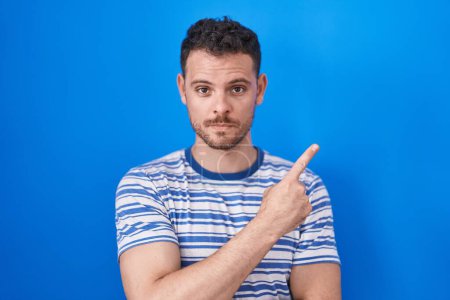 Téléchargez les photos : Jeune homme hispanique debout sur fond bleu pointant avec le doigt de la main sur le côté montrant la publicité, visage sérieux et calme - en image libre de droit