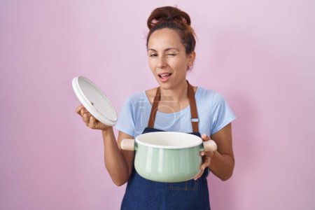 Téléchargez les photos : Brunette femme portant tablier tenant pot de cuisson clin d'oeil en regardant la caméra avec une expression sexy, gai et heureux visage. - en image libre de droit