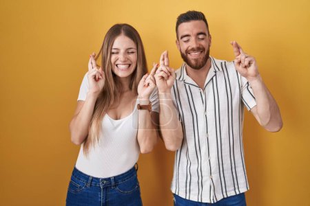 Photo for Young couple standing over yellow background gesturing finger crossed smiling with hope and eyes closed. luck and superstitious concept. - Royalty Free Image