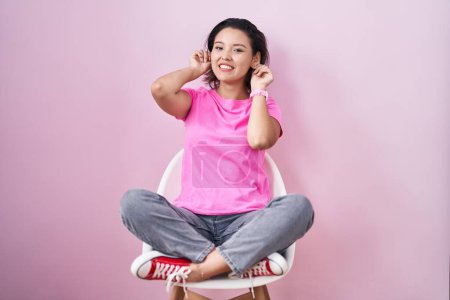 Foto de Mujer joven hispana sentada en silla sobre fondo rosa sonriendo tirando de las orejas con los dedos, gesto divertido. problema de audición - Imagen libre de derechos