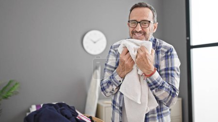 Téléchargez les photos : Homme d'âge moyen souriant confiant sentant les vêtements propres à la buanderie - en image libre de droit