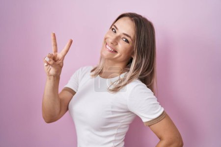 Téléchargez les photos : Femme caucasienne blonde debout sur fond rose souriant en regardant la caméra montrant les doigts faisant signe de victoire. numéro deux. - en image libre de droit