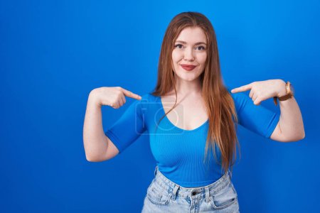 Photo for Redhead woman standing over blue background looking confident with smile on face, pointing oneself with fingers proud and happy. - Royalty Free Image