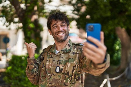 Foto de Joven hispano vistiendo uniforme camuflaje del ejército haciendo videollamada apuntando con el pulgar hacia el costado sonriendo feliz con la boca abierta - Imagen libre de derechos