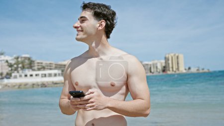 Téléchargez les photos : Jeune homme hispanique touriste souriant confiant en utilisant smartphone à la plage - en image libre de droit