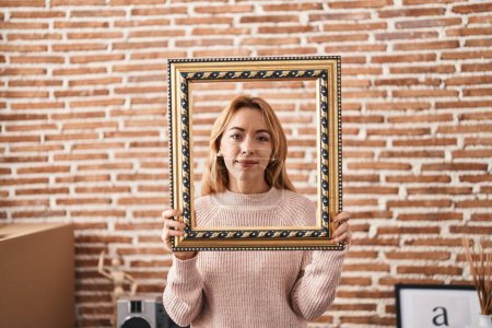 Photo for Hispanic woman holding empty frame relaxed with serious expression on face. simple and natural looking at the camera. - Royalty Free Image