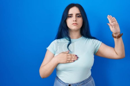 Photo for Young modern girl with blue hair standing over blue background swearing with hand on chest and open palm, making a loyalty promise oath - Royalty Free Image