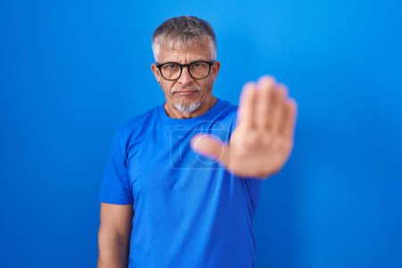 Photo for Hispanic man with grey hair standing over blue background doing stop sing with palm of the hand. warning expression with negative and serious gesture on the face. - Royalty Free Image