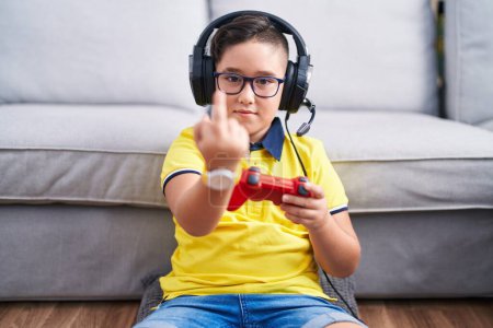 Foto de Young hispanic kid playing video game holding controller wearing headphones showing middle finger, impolite and rude fuck off expression - Imagen libre de derechos