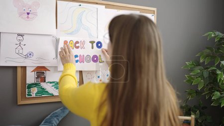 Téléchargez les photos : Jeune femme blonde enseignante préscolaire suspendue au papier de l'école sur carton-liège à la maternelle - en image libre de droit