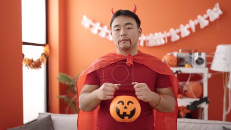 Foto de Young chinese man wearing devil costume holding halloween pumpkin basket at home - Imagen libre de derechos
